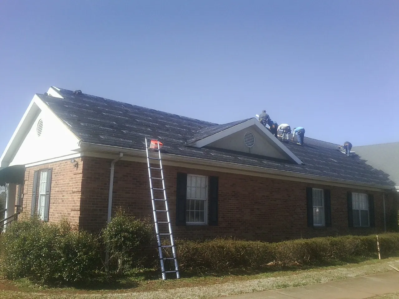 Crew tearing shingles off a brick ranch roof with a ladder set — Emergency Roof Repair work by our crew in Pfafftown