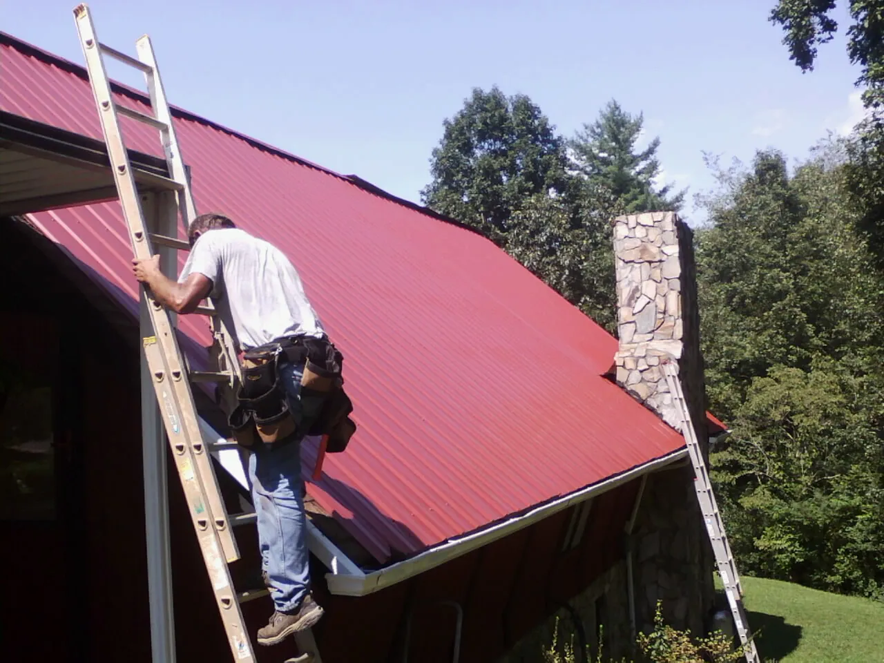 Roofer on a ladder beside a red metal roof and stone chimney — Soffit & Fascia Repair by Roofing Brothers in Pfafftown